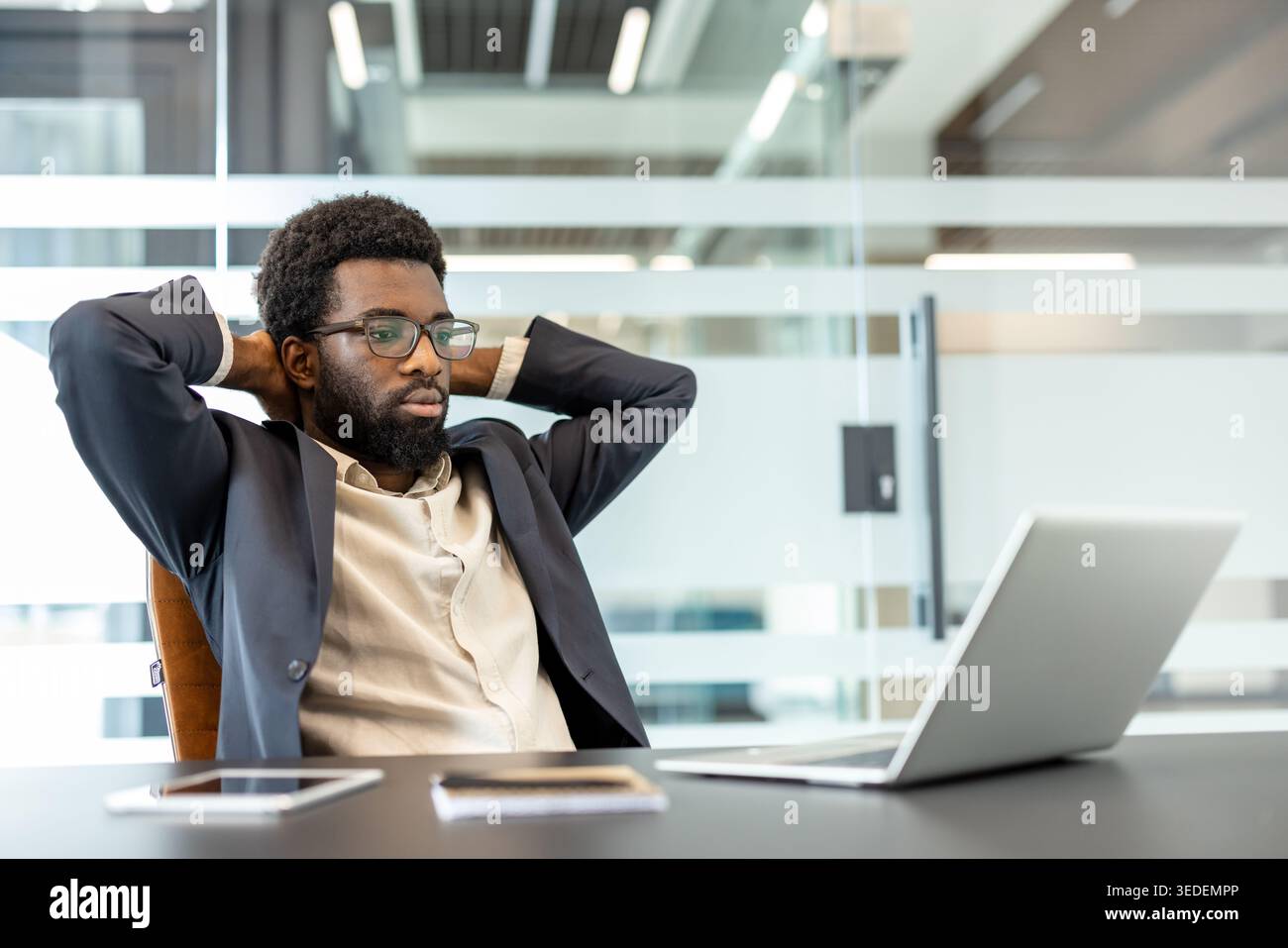 Un uomo d'affari concentrato in un ufficio moderno si piega con le mani dietro la testa, guardando il notebook mentre contempla strategia, la risoluzione dei problemi e i piani futuri con calma e sicurezza Foto Stock