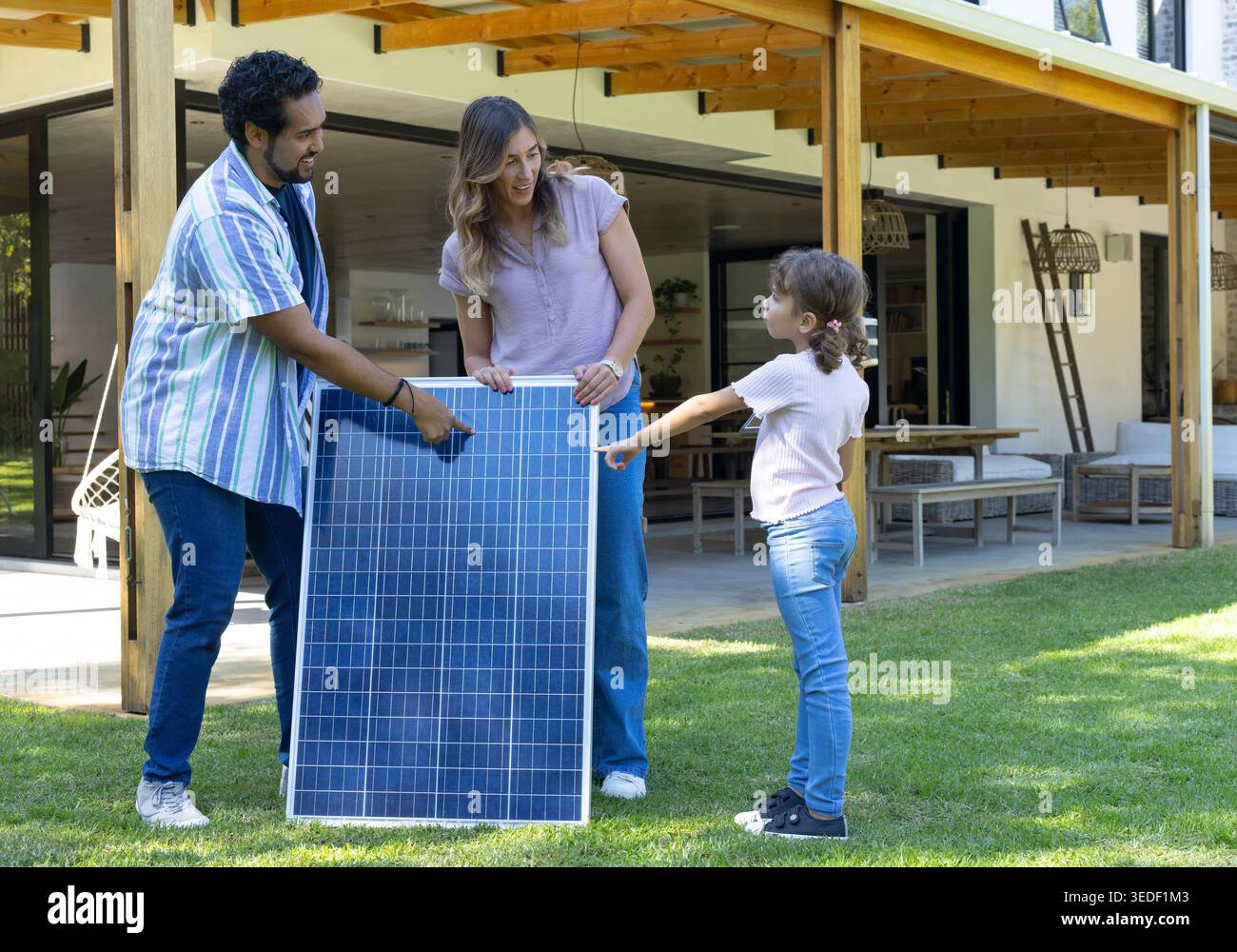 Famiglia con genitori e figlia che tengono il pannello solare verticale, che punta e parla sul prato Foto Stock