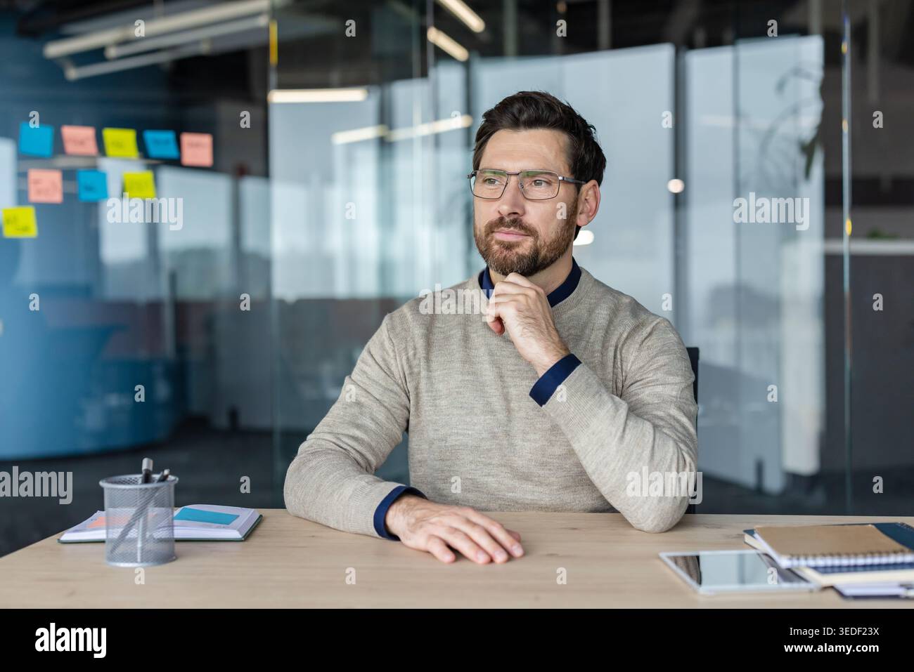 Uomo d'affari con la barba che indossa gli occhiali seduti alla scrivania, attento alla riflessione e alla pianificazione della strategia futura mentre lavora in un ambiente d'ufficio moderno Foto Stock