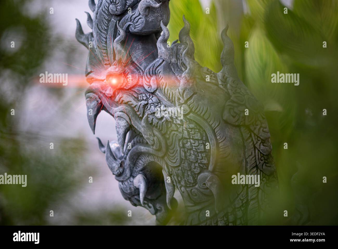 Primo piano di una scultura di drago intagliata con un'espressione feroce e occhi rossi brillanti, circondata da un lussureggiante fogliame verde, che mostra l'artigianato a. Foto Stock