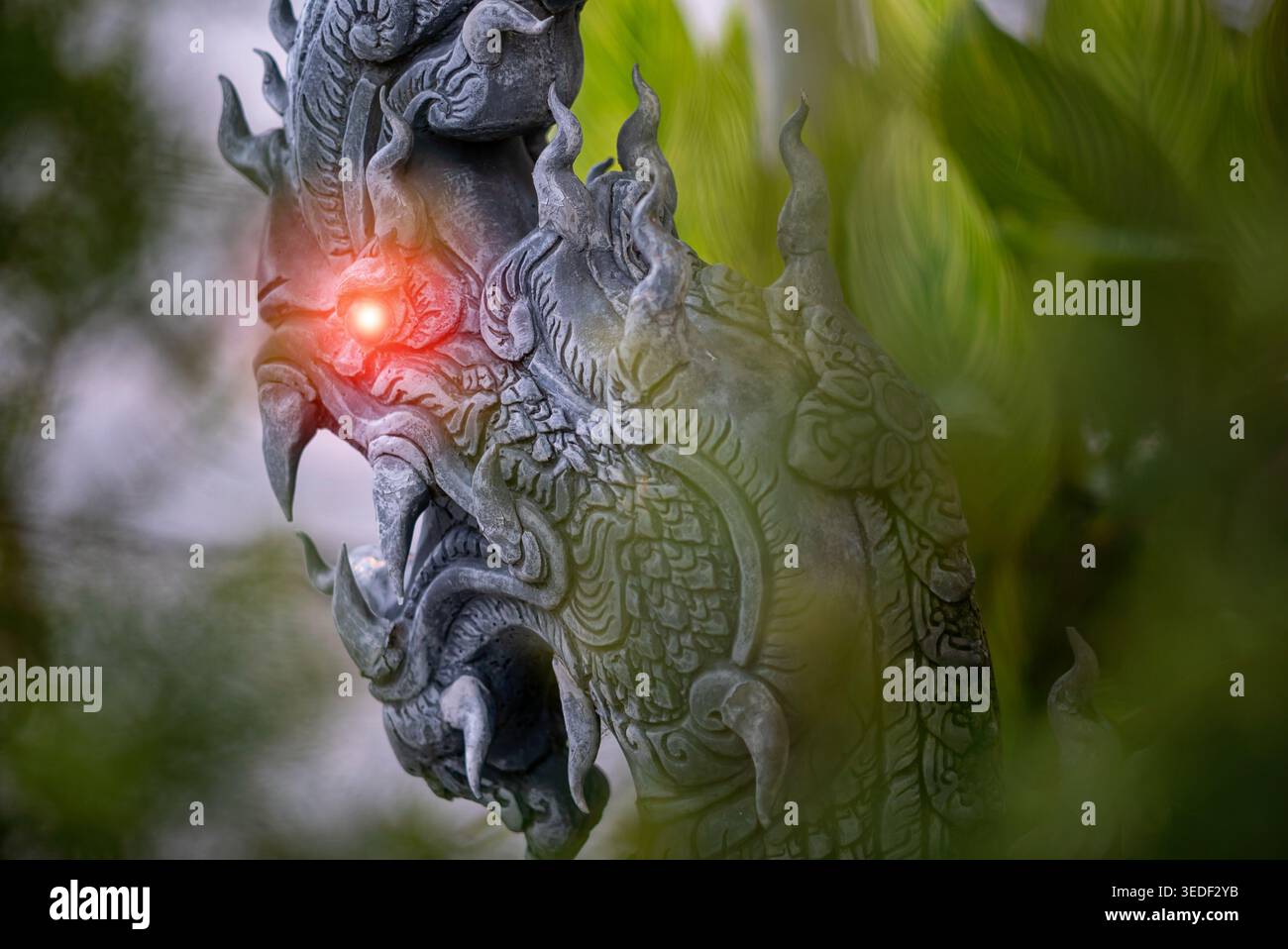 Un'accattivante scultura di drago in pietra caratterizzata da disegni intricati e occhi rossi brillanti. Sorge in mezzo a una lussureggiante vegetazione, evocando un'atmosfera mistica. Foto Stock