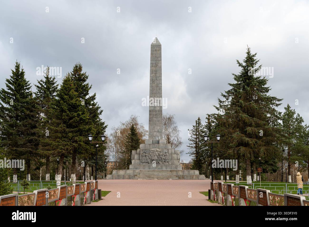 Viale simmetrico che conduce all'Obelisco centrale, il corridore mattutino attraversa la passeggiata in mattoni, le file Evergreen creano l'effetto tunnel, le panchine vuote Foto Stock