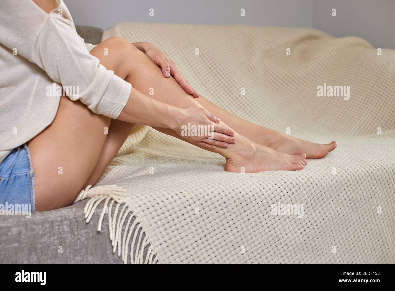 Donna seduta su un divano massaggiando lo stinco e il polpaccio per alleviare dolore muscolare, crampi o stanchezza dopo l'esercizio fisico Foto Stock