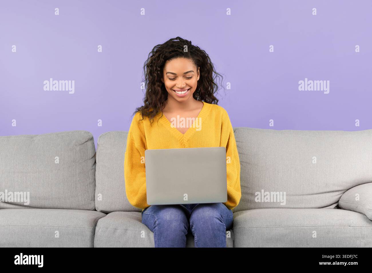 La donna si siede su un divano con un notebook e sorride mentre guarda lo schermo Foto Stock