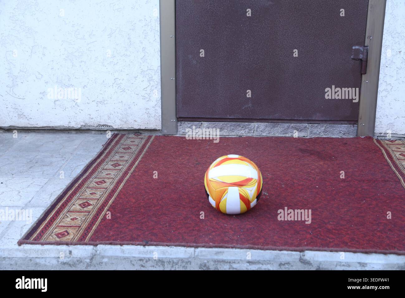 Una palla da calcio si trova su un tappetino rosso di fronte a una porta chiusa. L'ambientazione è durante il giorno e non ci sono persone in vista. La scena mostra un momento tranquillo in un quartiere Foto Stock