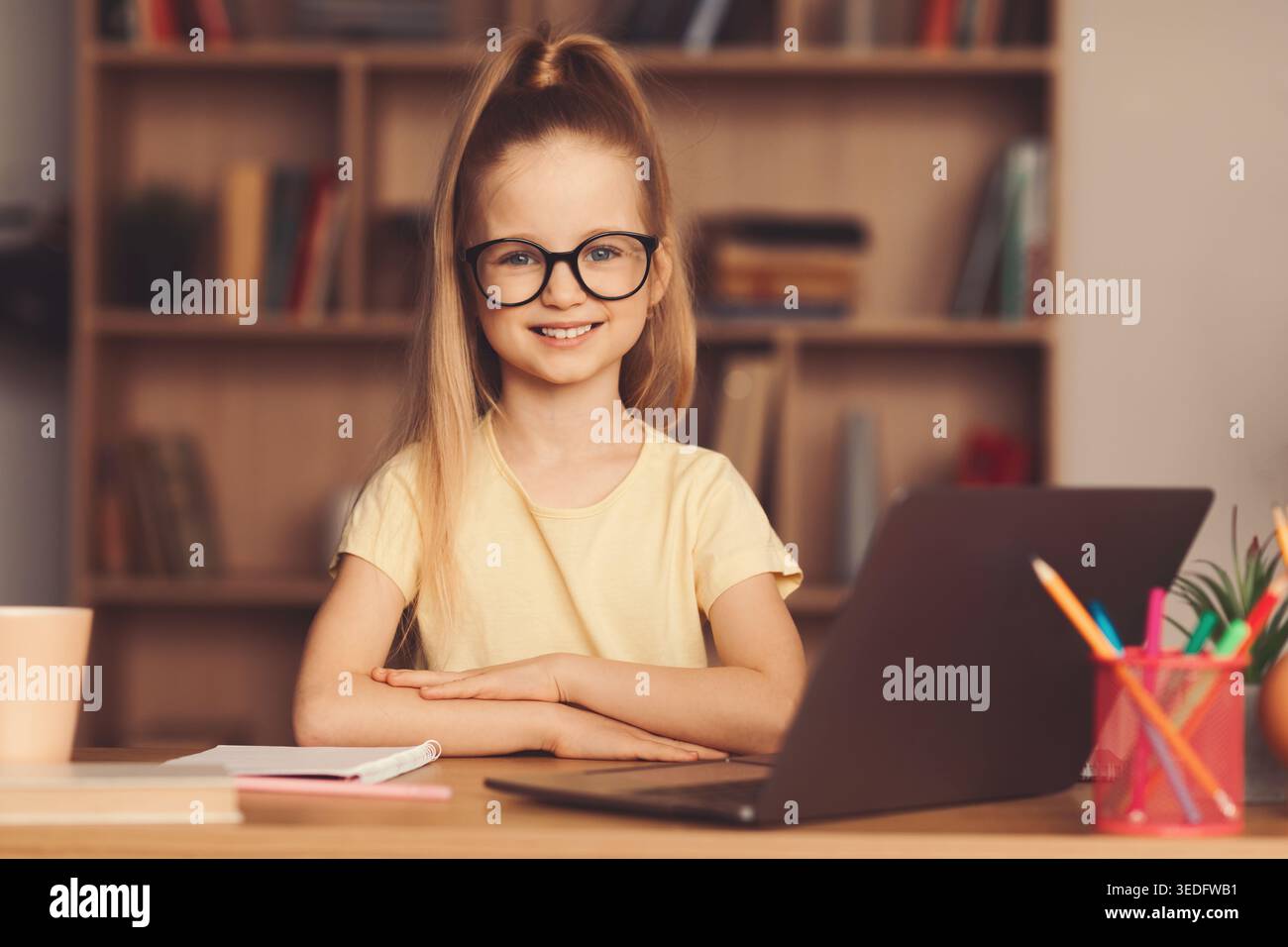 La ragazza con gli occhiali sorride mentre si siede alla scrivania con laptop e materiale scolastico in una sala studio Foto Stock