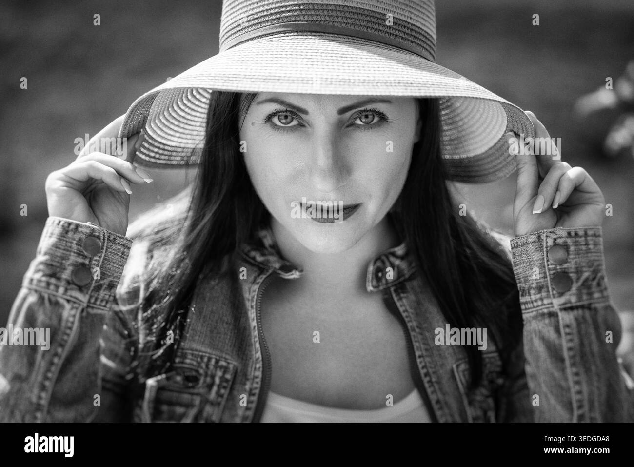 Ritratto di donna che indossa un cappello di paglia estivo e una giacca in denim. Fotografia in bianco e nero in stile vintage con pellicola granulosa. Elegante signora che guarda Foto Stock