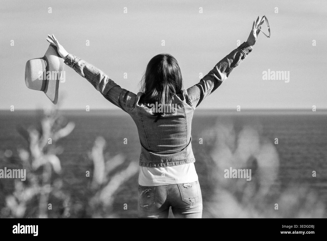 Vista posteriore di una donna in piedi con le braccia sollevate che regge un cappello di paglia che guarda al mare. Vista posteriore della signora che gode della libertà. Foto in bianco e nero in stile vintage Foto Stock