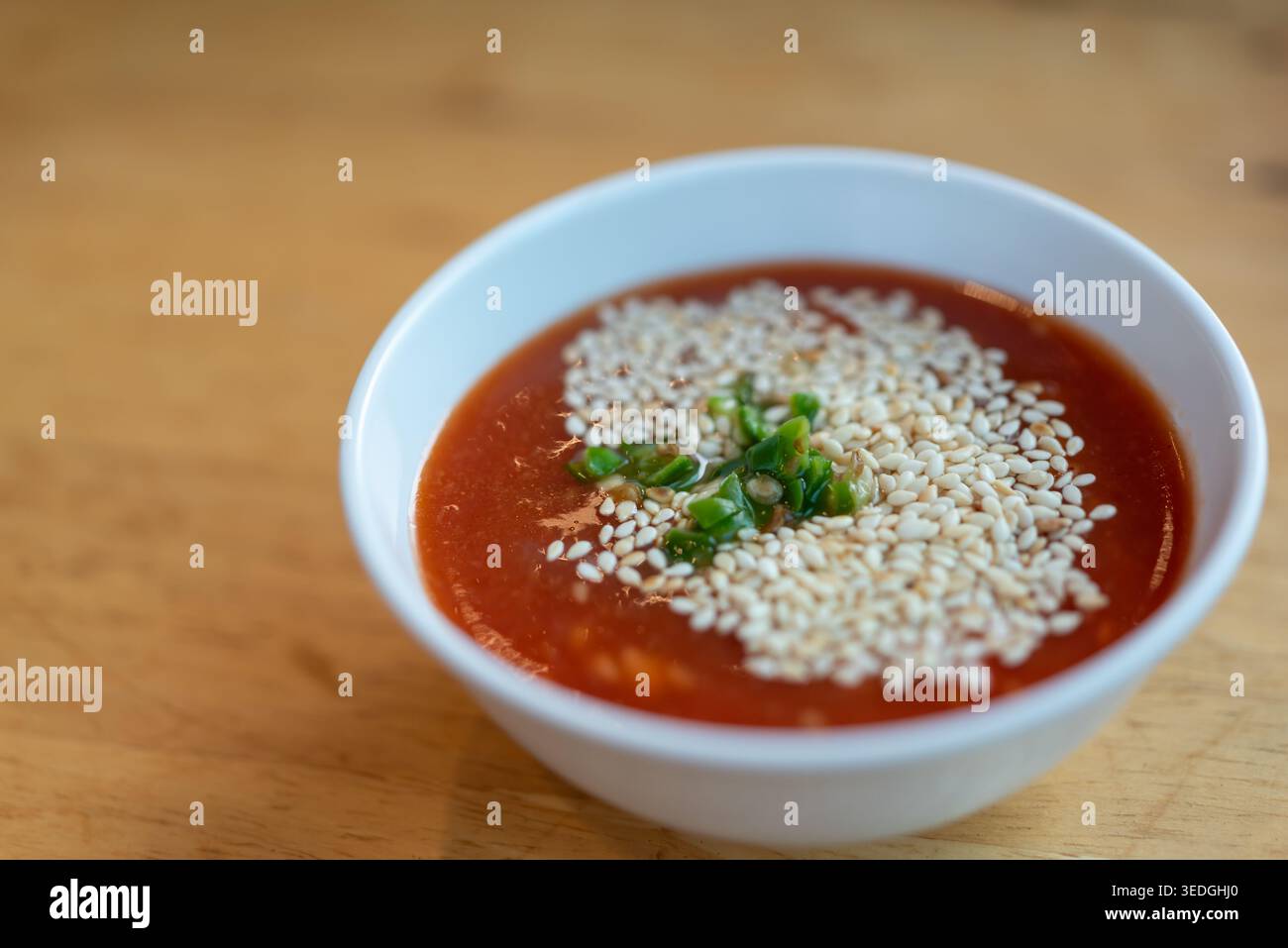 Primo piano di salsa rossa piccante con semi di sesamo e peperoncino verde tritato in una ciotola bianca su un tavolo di legno dall'aroma ricco di salsa asiatica Foto Stock