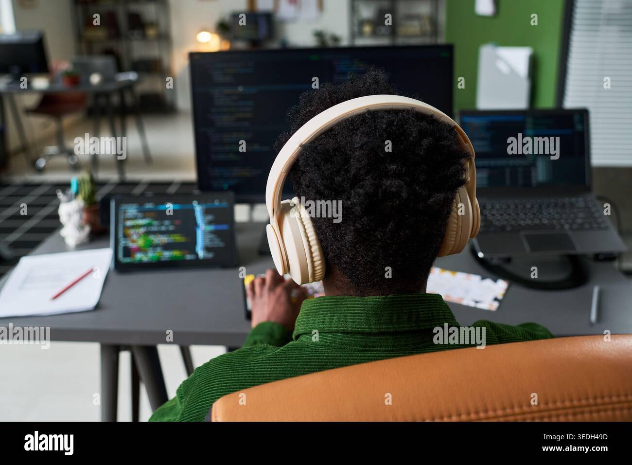 Vista posteriore del giovane uomo nero che indossa le cuffie, codifica su più schermi alla scrivania, che rappresenta lo sviluppo del software e il lavoro in remoto. Ideale per carriere tecnologiche o servizi IT Foto Stock