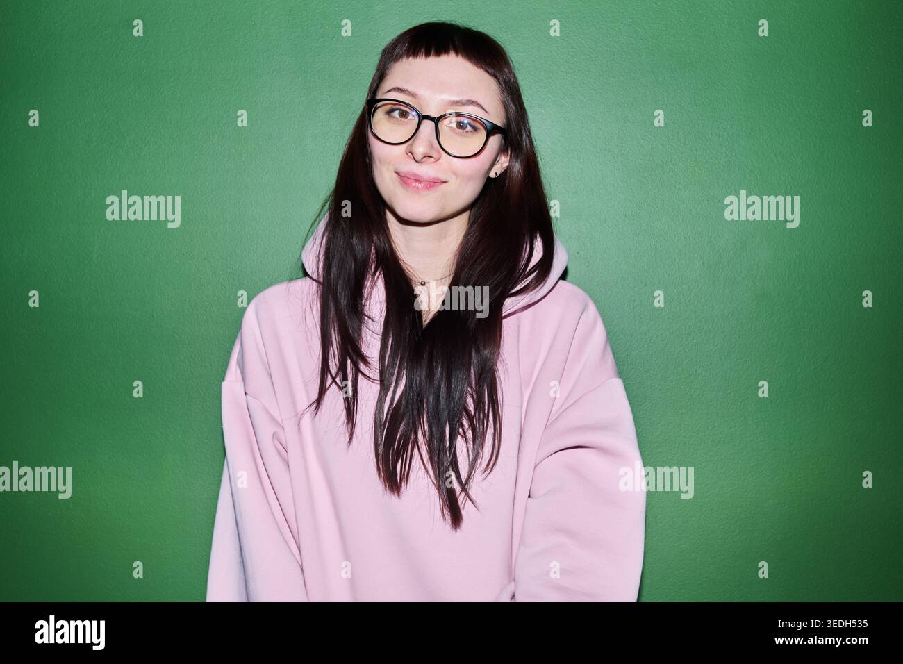 Ritratto di una giovane donna caucasica adulta che indossa gli occhiali, sorridendo alla fotocamera su sfondo verde, trasmettendo fiducia e individualità. Utile per il branding lifestyle o per gli annunci di occhiali Foto Stock