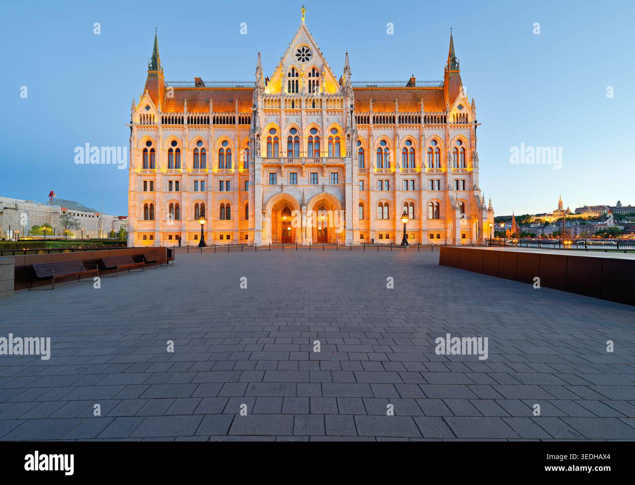 I visitatori camminano intorno all'edificio del Parlamento di Budapest mentre la luce del tramonto proietta un caldo bagliore sulla struttura e sull'acqua nelle vicinanze. Foto Stock
