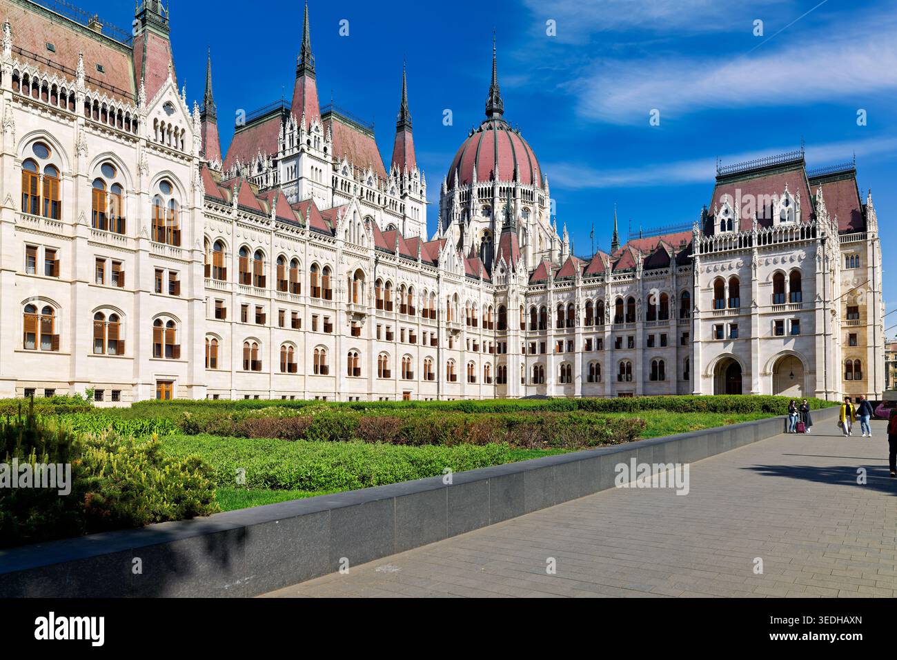 La gente cammina lungo il sentiero vicino all'edificio del Parlamento a Budapest. Il sole splende sulla struttura storica e sullo spazio verde. Foto Stock