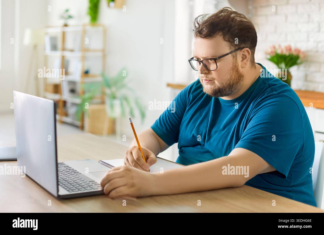 Uomo in sovrappeso che prende appunti mentre studia online a casa con un portatile sulla scrivania Foto Stock