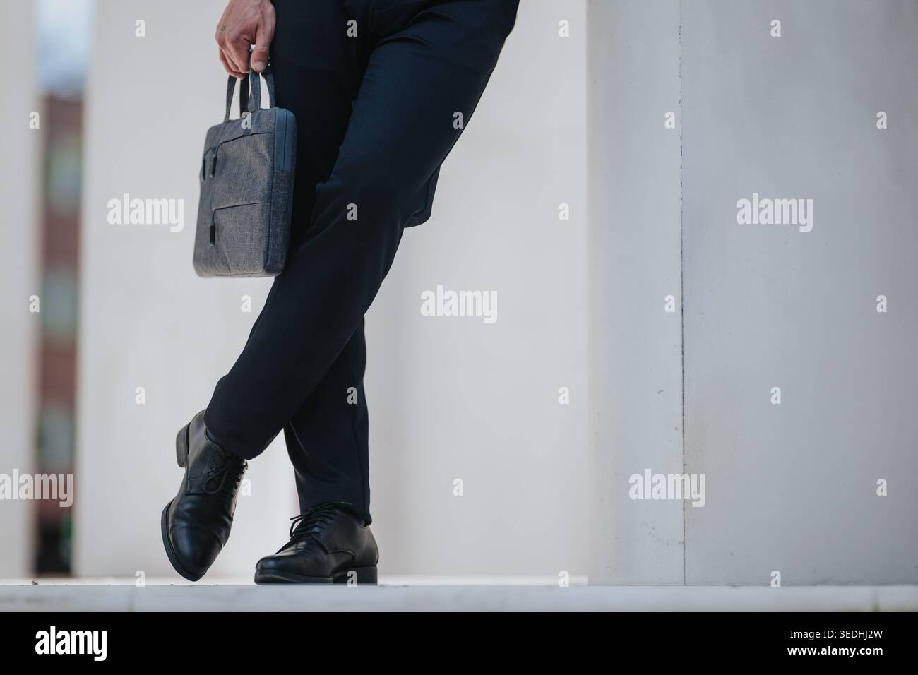 Uomo d'affari appoggiato a una colonna con gambe incrociate che reggono una valigetta grigia e scarpe in pelle Foto Stock