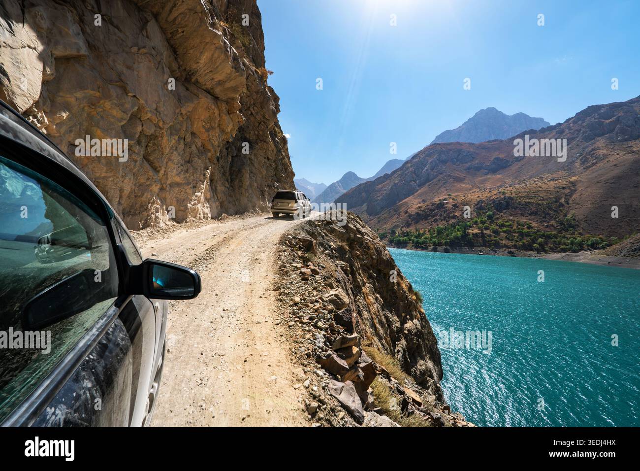 Un'auto guida su una pericolosa strada di montagna con scogliere sulla sinistra e una discesa a picco in un lago di montagna sulla destra. Strada panoramica estrema in Tajikista Foto Stock