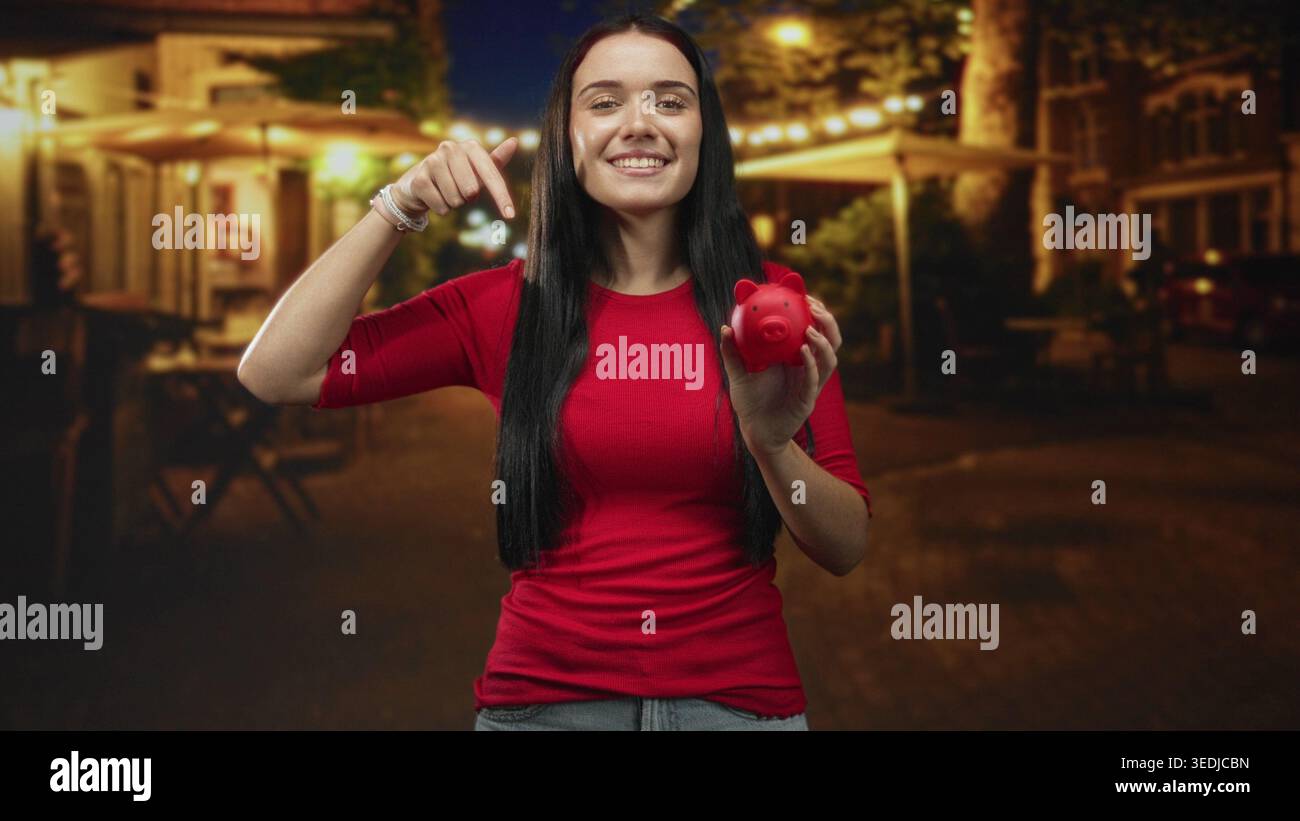 Una giovane donna in camicia rossa punta il dito verso il salvadanaio mentre lo tiene in una strada notturna acciottolata; ottimismo finanziario. Foto Stock