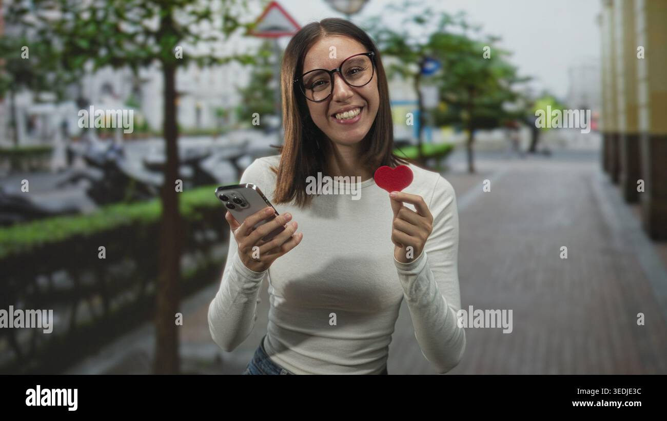 Donna in occhiali con un piccolo cuore rosso e sorridente mentre legge un messaggio sul suo smartphone in jeans casual su una strada della città costeggiata da alberi; ro Foto Stock