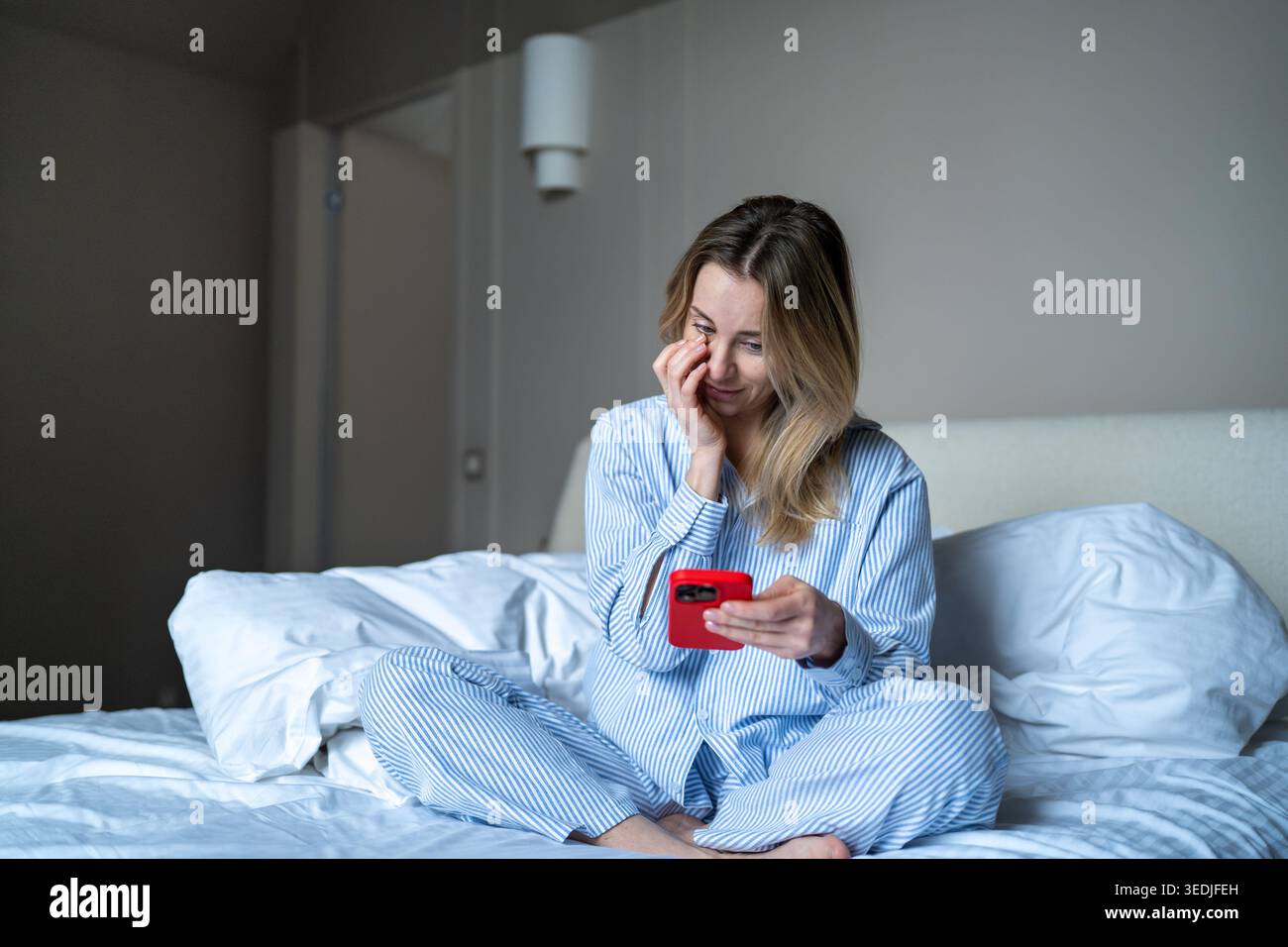 Mattina online con il telefono. Una donna contenta usa lo smartphone seduto a letto scorrendo le ragnatele dopo il risveglio Foto Stock