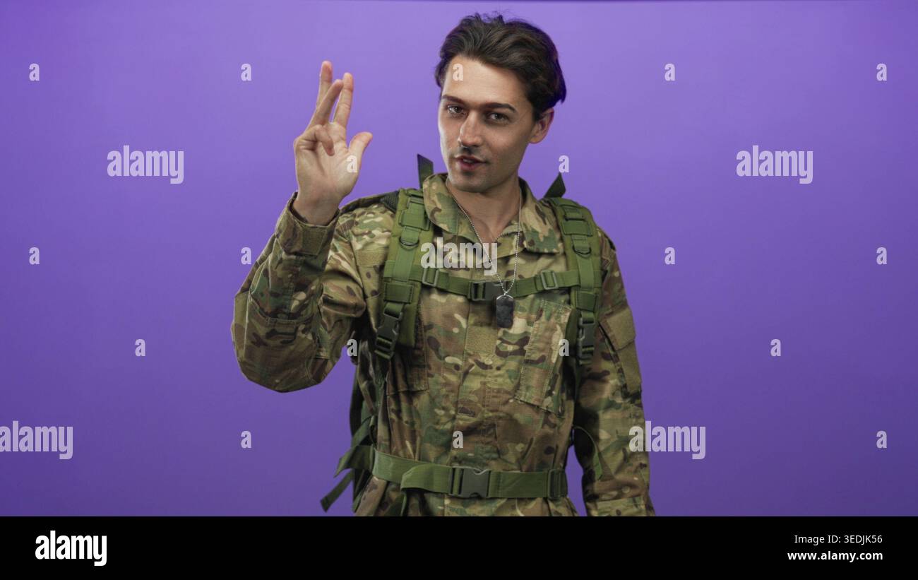 Uomo in uniforme militare mimetica con collana dogtag e zaino tattico solleva due dita in un gesto di pistola vicino al viso in studio viola ba Foto Stock