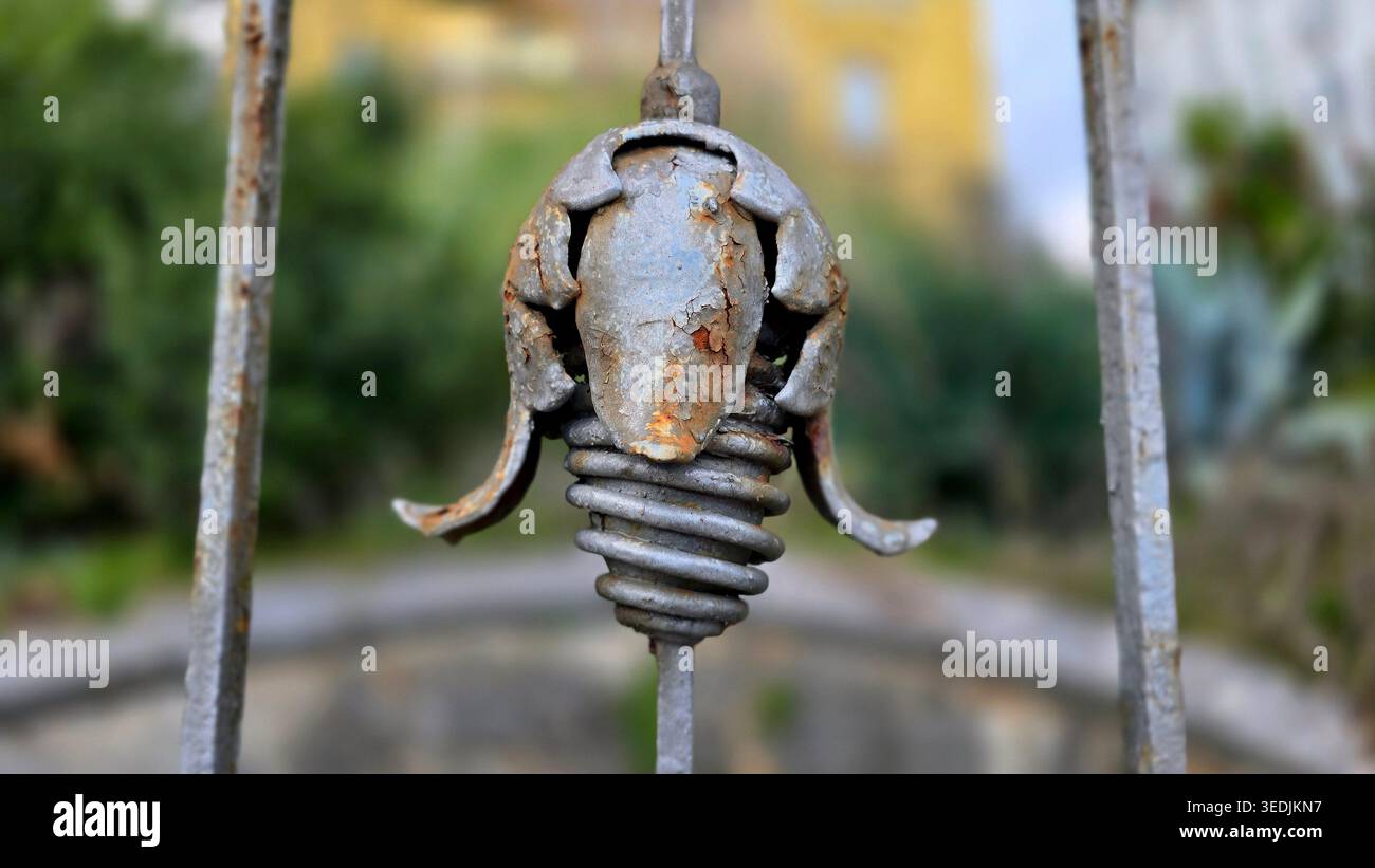 Una foto dettagliata di un ornamento metallico grigio con forme a spirale e floreali su un vecchio cancello. Lo sfondo presenta una morbida vegetazione sfocata Foto Stock