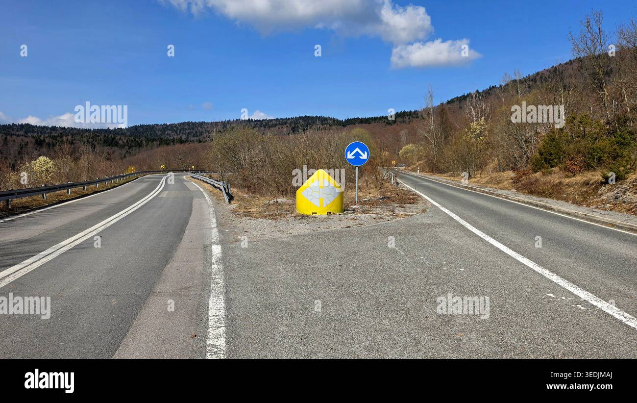 Una strada asfaltata si divide in due direzioni sotto un cielo blu brillante con nuvole bianche. La scena rurale presenta barriere di sicurezza e indicatori di traffico Foto Stock