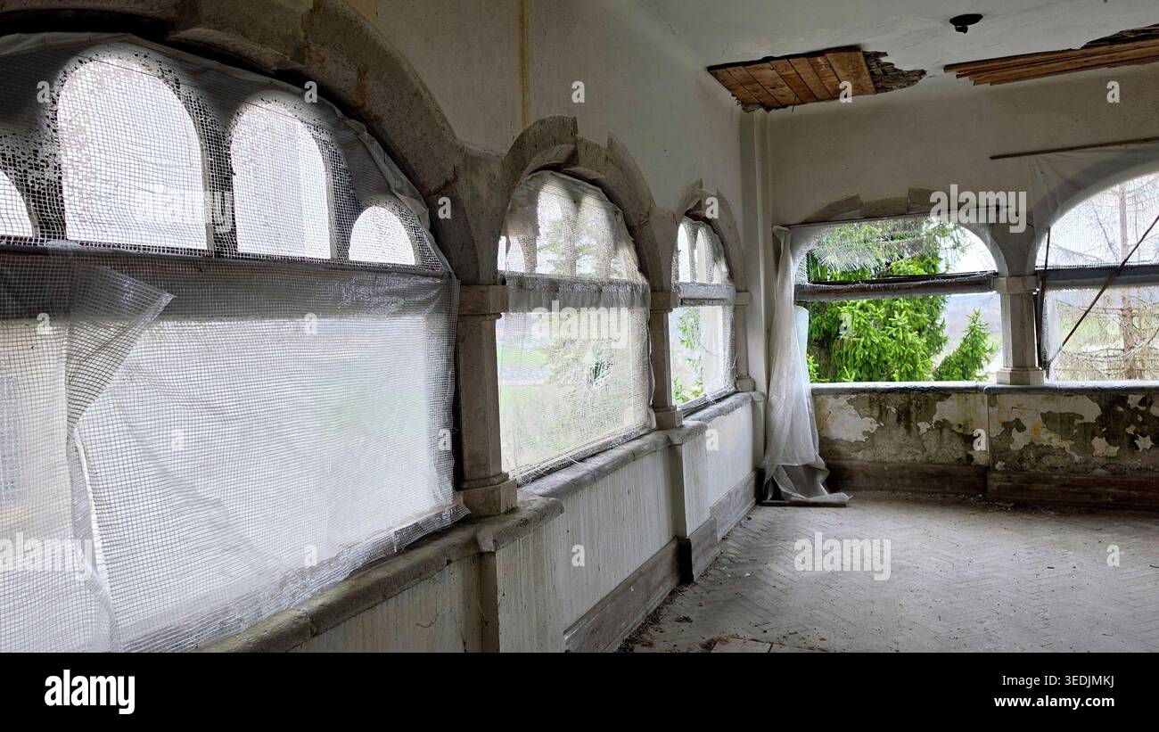 Una lunga stanza in una tenuta trascurata presenta una serie di finestre ad arco in pietra protette da plastica traslucida. Lo spazio mostra muri che decadono, un danno Foto Stock