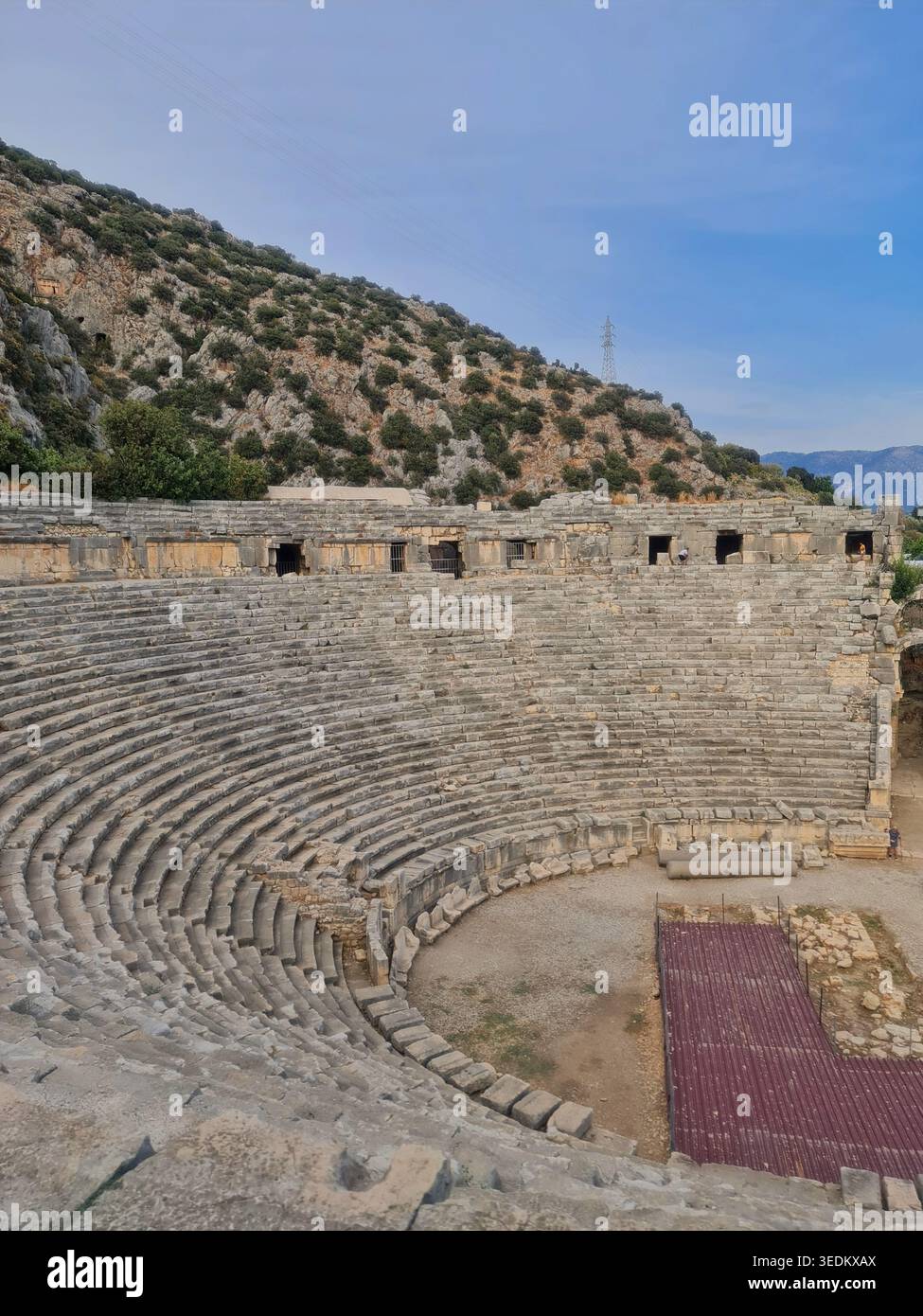 Storico anfiteatro greco-romano con posti a sedere in pietra semicircolare e rovine intemprate adagiate su una collina rocciosa mediterranea sotto un chiaro sk blu Foto Stock