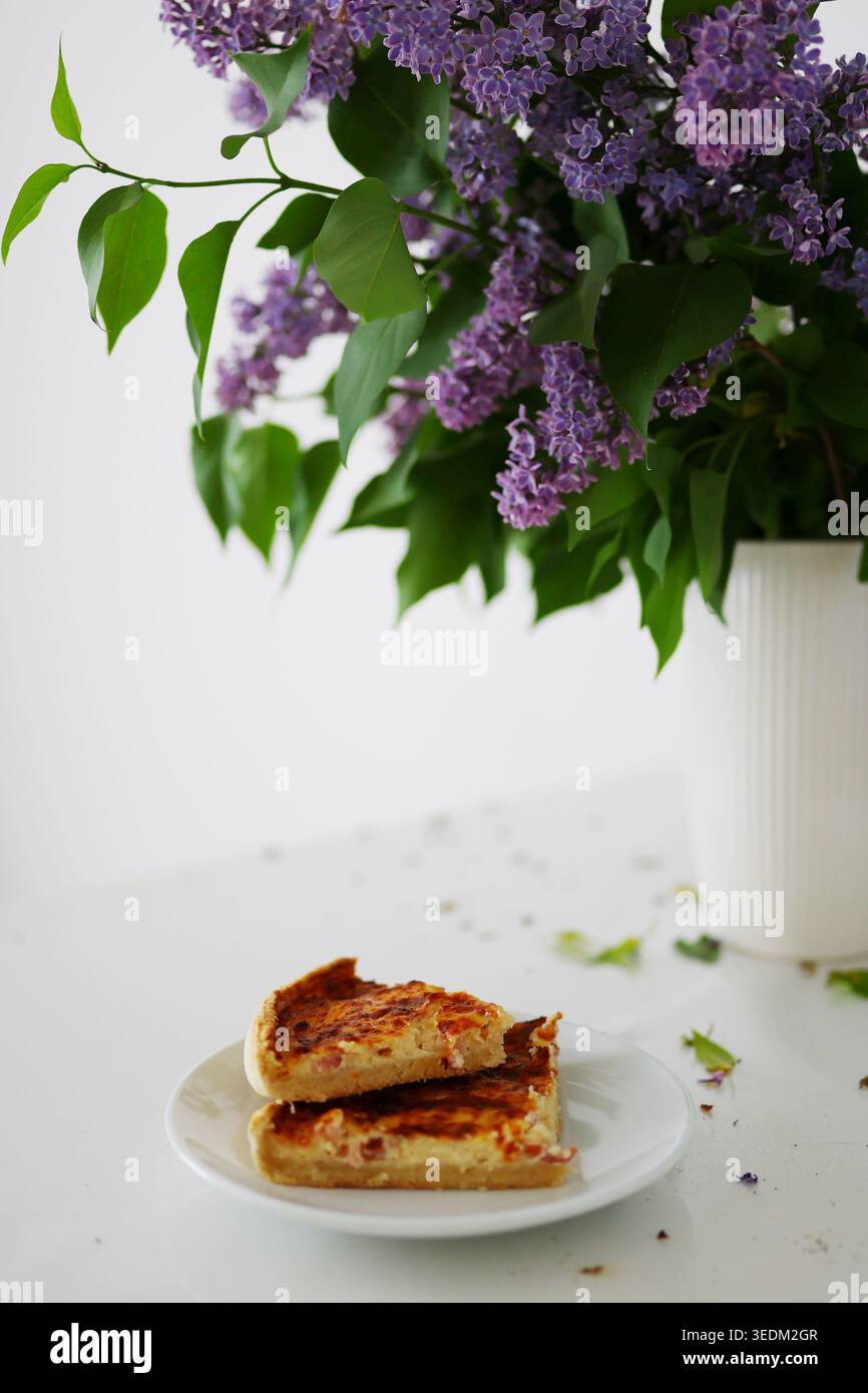Natura morta primaverile con crostata fatta in casa e fresco bouquet lilla su sfondo bianco, cucina accogliente e atmosfera stagionale. Foto Stock