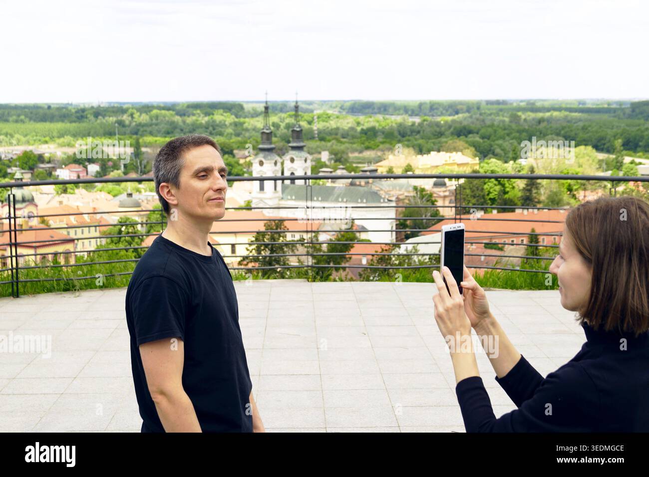 Donna che punta la fotocamera dello smartphone all'uomo con un paesaggio urbano sfocato di Sremski Karlovci. Documentazione di viaggio curata dai partner e raccolta di memorie esperienziali Foto Stock