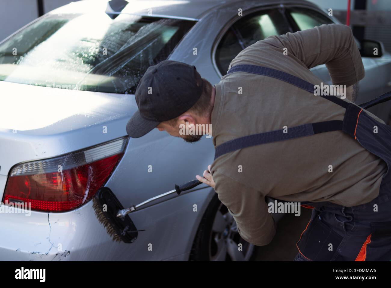 Uomo in grembiule e berretto strofinando con cautela il paraurti posteriore e le luci di posizione posteriori di una berlina argentata con una spazzola a manico lungo e suds saponati durante una CA all'aperto Foto Stock