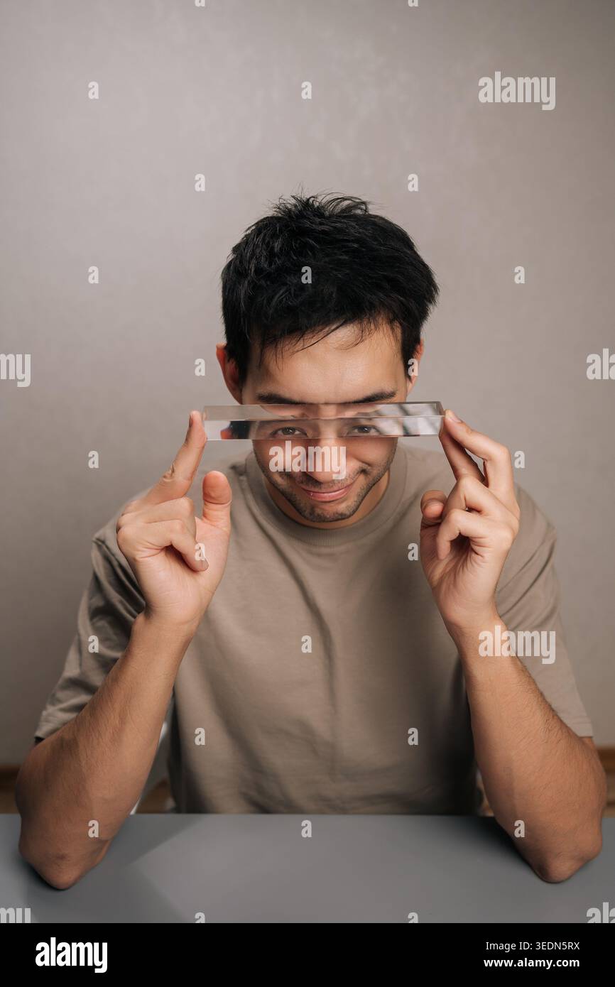 Ripresa verticale di un uomo sorridente che tiene il prisma di vetro davanti agli occhi, che mostra la rifrazione della luce e la distorsione ottica, esplorando giocosamente la visuale Foto Stock