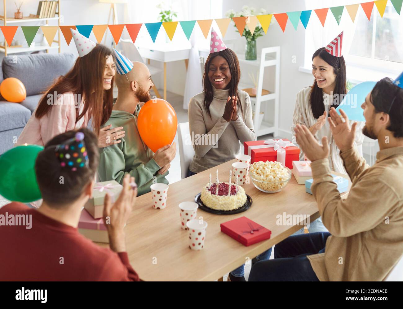 Un gruppo di giovani amici felici e diversi si riunì intorno al tavolo per festeggiare il compleanno e godersi la festa. Foto Stock