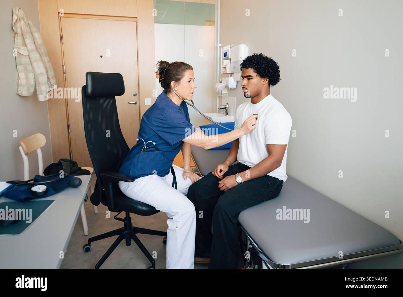 Donna medico professionista che ascolta il battito cardiaco del giovane uomo attraverso lo stetoscopio in sala d'esame all'ospedale Foto Stock