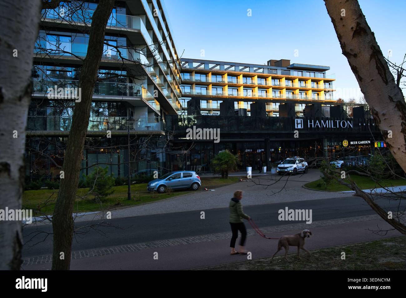 Facciata moderna dell'hotel Hamilton al tramonto, con cane pedonale e auto parcheggiate, la città costiera di Świnoujście. Foto Stock