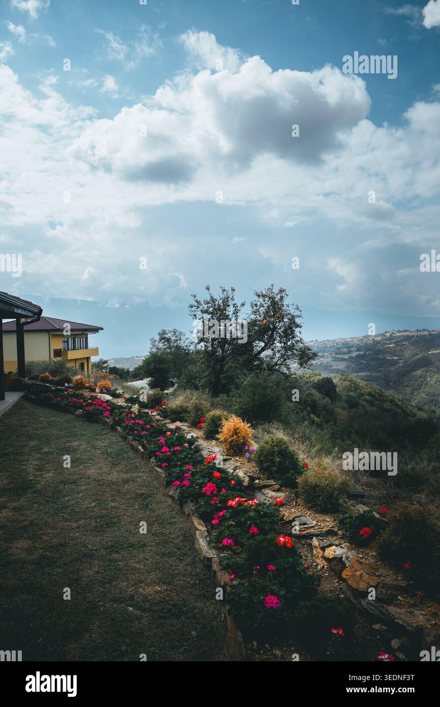 Petrich, Bulgaria - 09 26 2024: Un vivace giardino fiorito fiancheggiato da un confine in pietra accanto a una casa di campagna, che si affaccia su colline e montagne ondulate Foto Stock