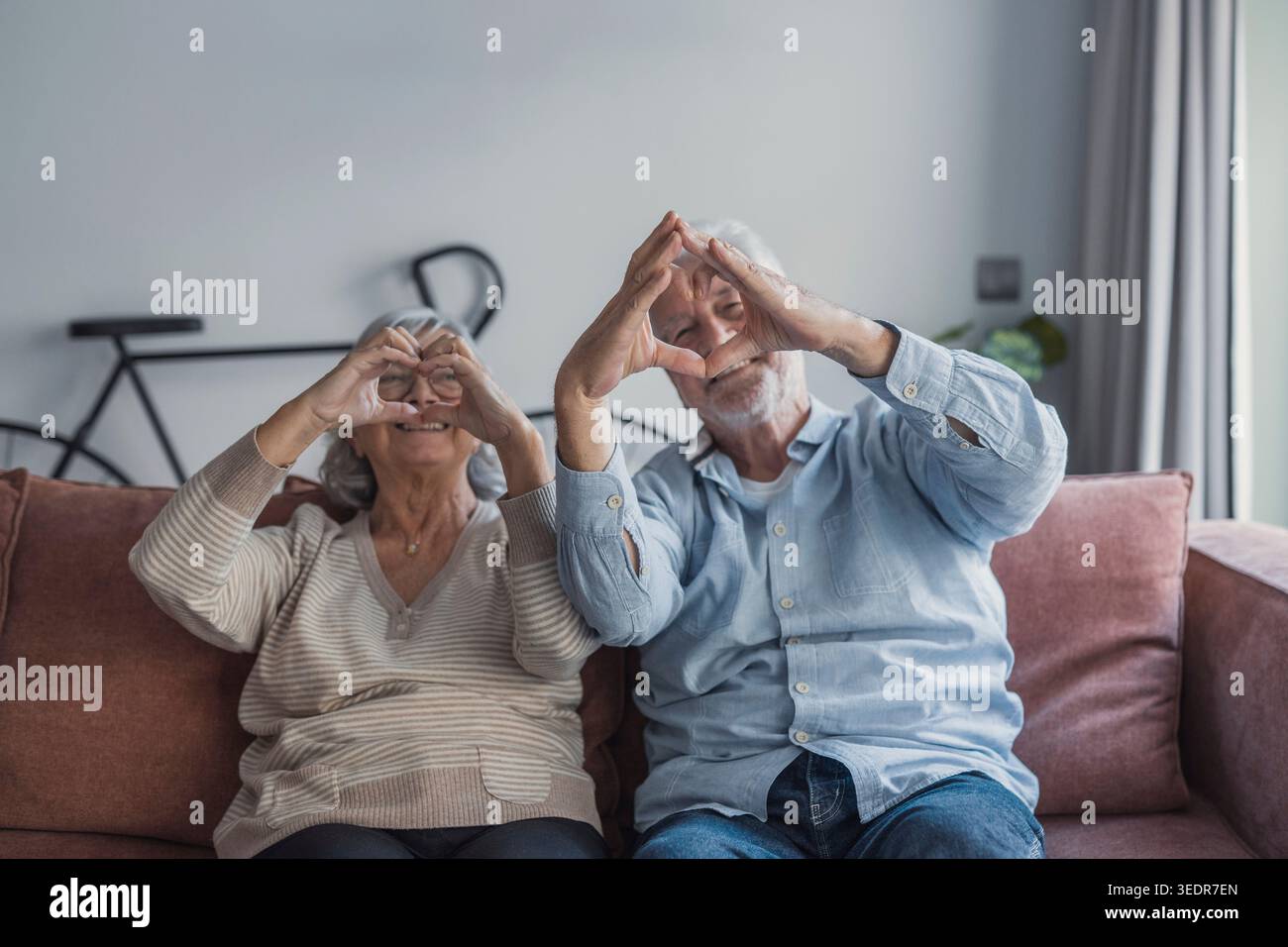 Una coppia anziana amorevole sta insieme in casa facendo un gesto cardiaco, esprimendo compassione, impegno e felicità condivisa in un ritratto intimo ed emotivo Foto Stock
