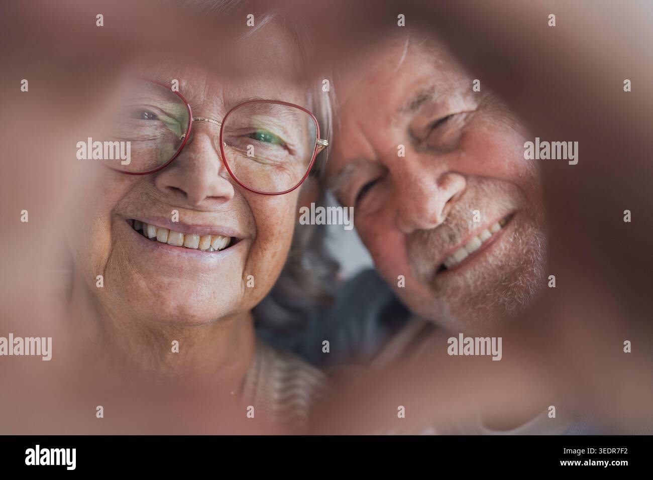 Una coppia in pensione felice posa in casa facendo un gesto cardiaco con le dita, sorridendo alla fotocamera ed esprimendo amore, sincerità e un forte legame emotivo in un ritratto caldo Foto Stock