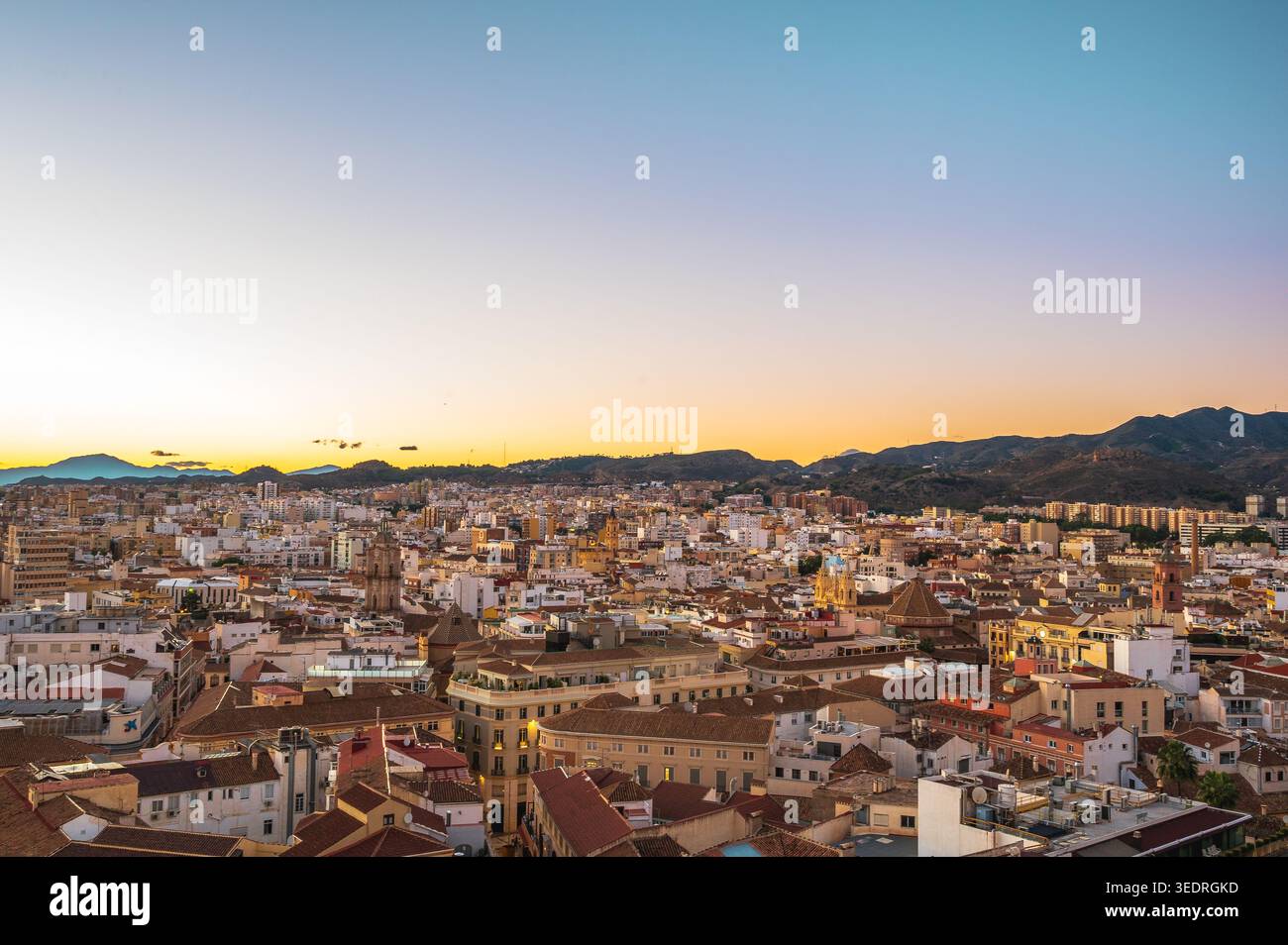 Ampia vista panoramica sulla città di Malaga, in Spagna, che mostra i fitti edifici urbani e i tetti con le colline circostanti sotto un colorato cielo al tramonto. Il Foto Stock