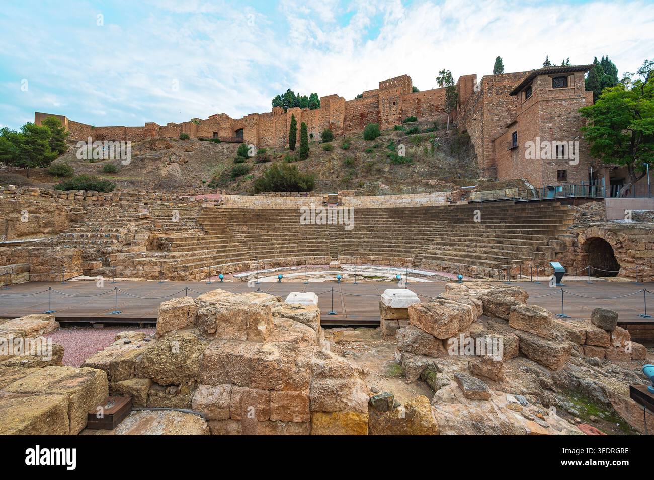 Antico teatro romano a Malaga, Spagna, con posti a sedere concentrici in pietra, palco in legno e rovine di roccia in primo piano situate sotto le mura della fortezza di Alcazaba Foto Stock