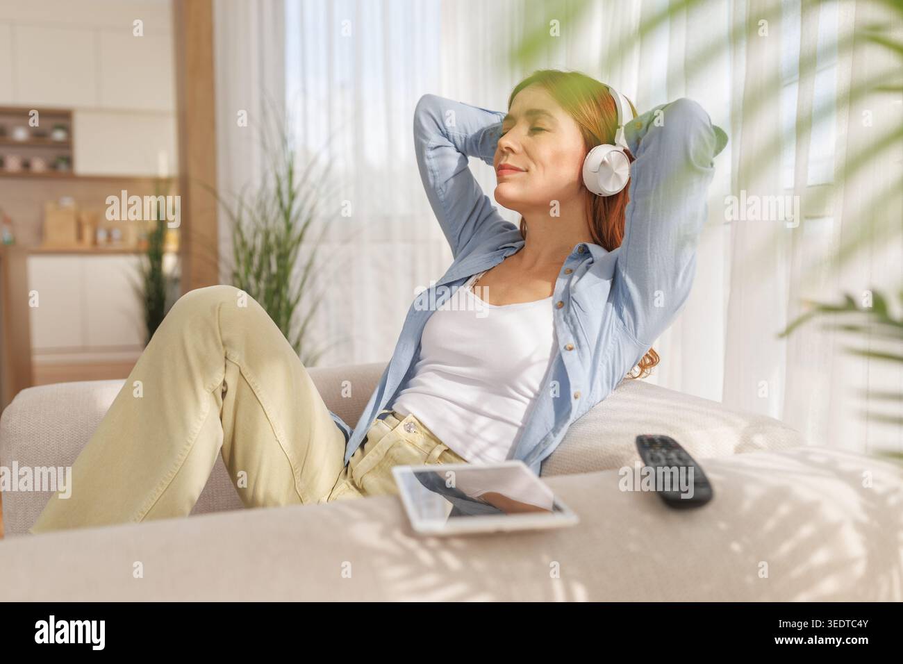 Una donna si siede su un divano in salotto e ascolta musica. Si piega indietro con gli occhi chiusi. Una tavoletta è accanto a lei sul divano. Foto Stock