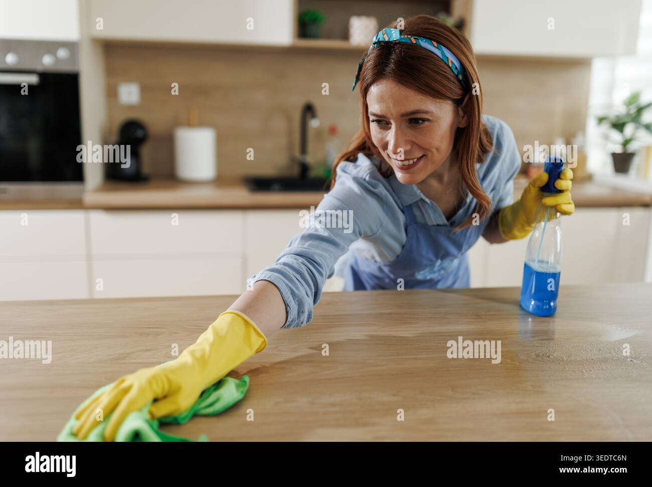 Una donna con guanti gialli utilizza un flacone spray e un panno per pulire la superficie della cucina. Si concentra sul suo compito in una cucina luminosa Foto Stock