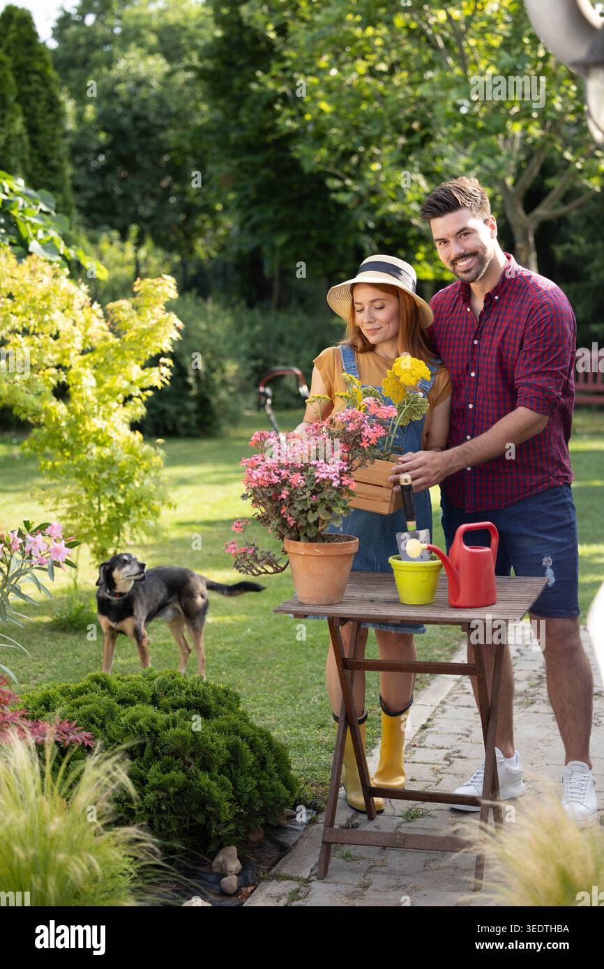 Una coppia si trova accanto a un tavolo piantando fiori in pentole mentre si gode una giornata di sole nel loro giardino. Un cane li guarda da lontano Foto Stock