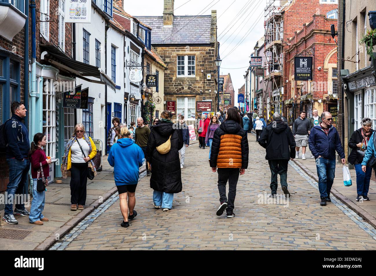 Trafficata strada acciottolata a Whitby, North Yorkshire, fiancheggiata da tradizionali edifici in mattoni e pietra che ospitano negozi e aziende. La gente cammina lungo la n Foto Stock