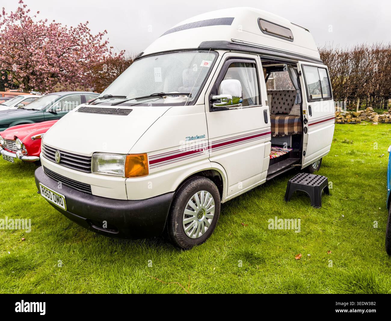 Camper Volkswagen T4 d'epoca bianca con un tetto alto parcheggiato su erba verde con la porta laterale aperta e fiori di ciliegio sullo sfondo Foto Stock