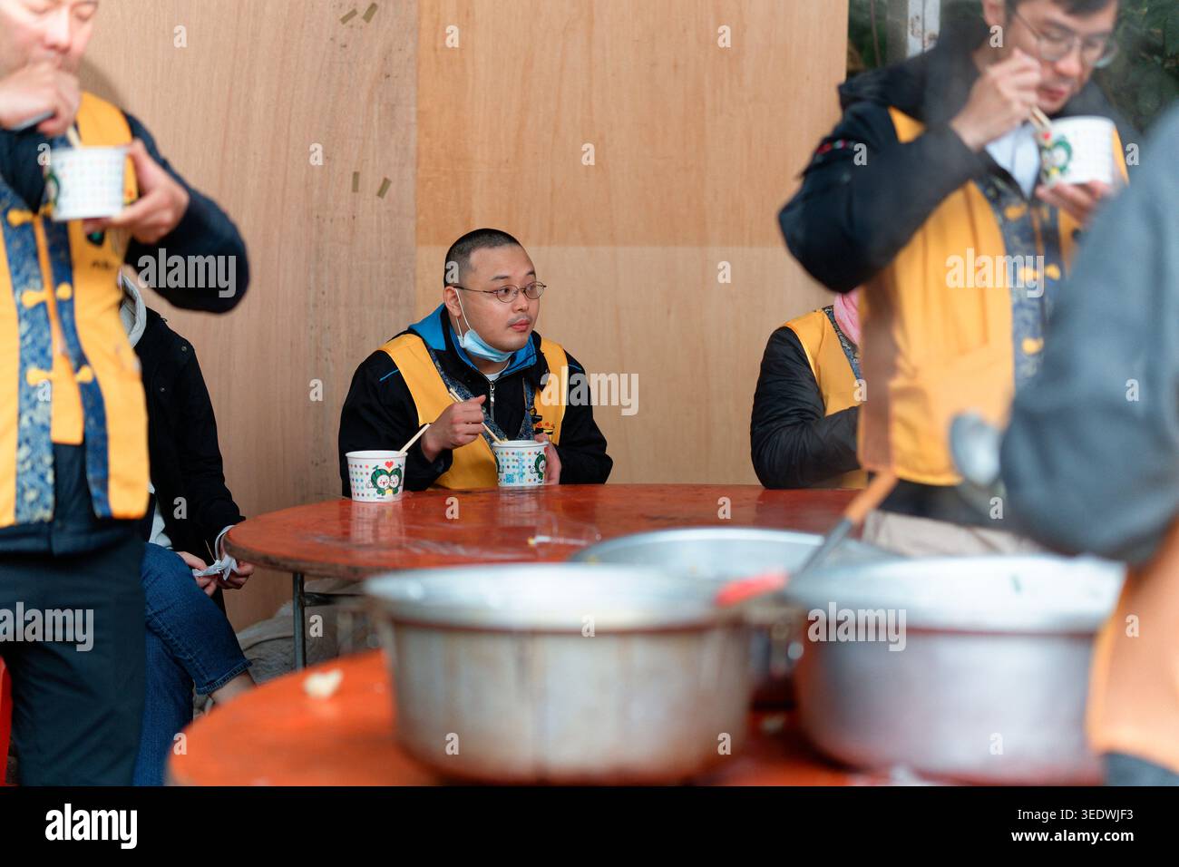 Taipei, Taiwan - 29 marzo 2025: I volontari del Tempio in gilet gialli mangiano da tazze di carta ai tavoli pieghevoli rossi durante un banchetto comune all'aperto. Foto Stock