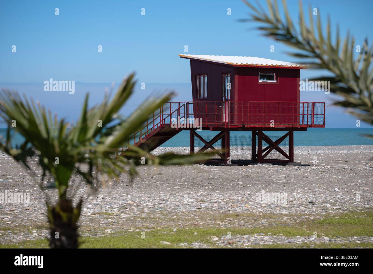 Capanna rossa di sicurezza in legno su palafitte situata su una spiaggia sassosa con foglie di palma in primo piano Foto Stock