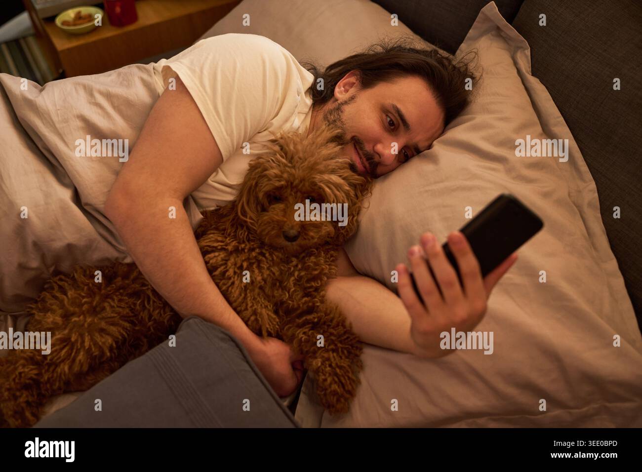 Giovane uomo caucasico disteso a letto, coccolando il cane e utilizzando lo smartphone per videochiamate o social media. Mostra compagnia domestica e stile di vita digitale per la cura degli animali domestici o l'app marketing Foto Stock