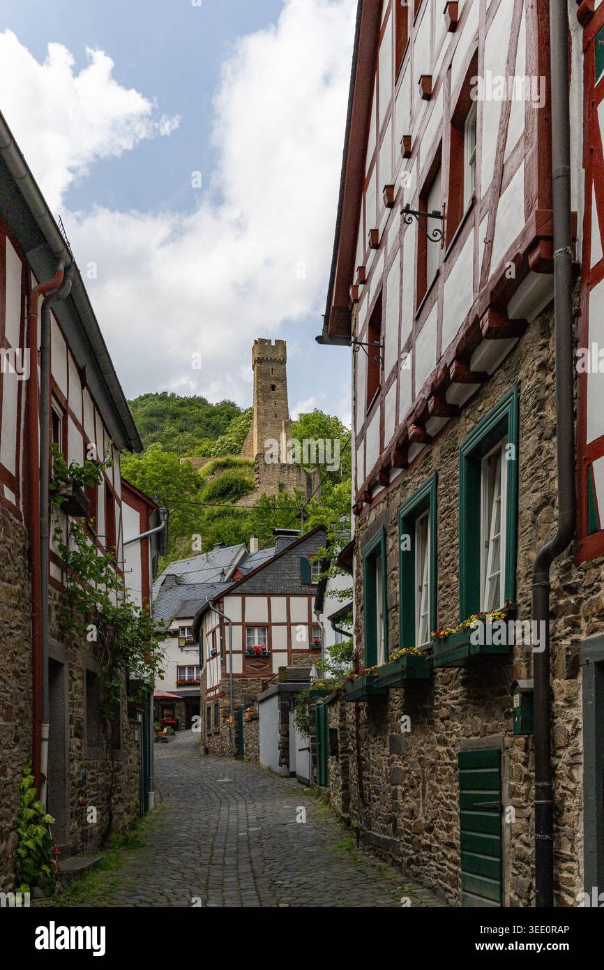 Veduta di una stretta strada acciottolata fiancheggiata da tradizionali case a graticcio che conducono verso una torre medievale del castello su una lussureggiante collina verde sotto a. Foto Stock