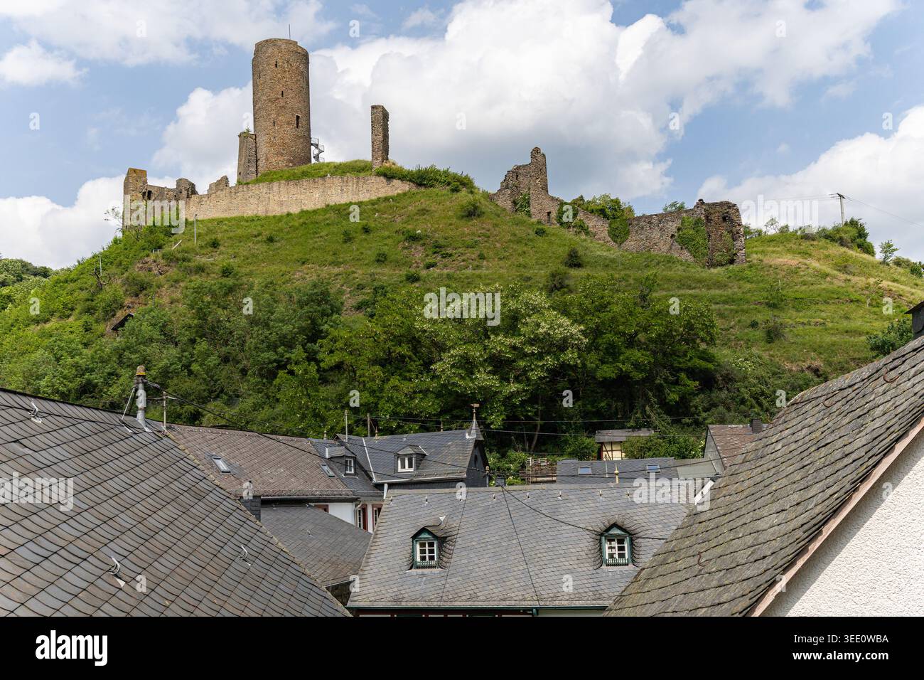 Vista delle storiche rovine del castello di Monreal su una lussureggiante collina verde sopra i tradizionali tetti di ardesia sotto un cielo nuvoloso a Monreal, Rheinland-Pfalz, in tedesco Foto Stock